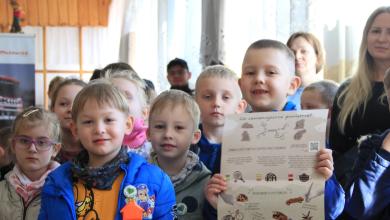 Na zdjęciu widzimy grupę dzieci biorących udział w akcji edukacyjnej w szkole.  Na pierwszym planie stoi trzech chłopców, którzy trzymają plakaty o tematyce ekologicznej.  Jeden z plakatów, trzymany przez chłopca po lewej stronie, zatytułowany jest „Jak dbać o Ziemię?” i prezentuje kolorową grafikę z planetą Ziemia oraz hasłami promującymi aspekty działalności proekologicznej, takimi jak oszczędzanie wody, segregowanie śmieci czy oszczędzanie energii.  Chłopiec po prawej stronie trzyma plakat zatytułowany „